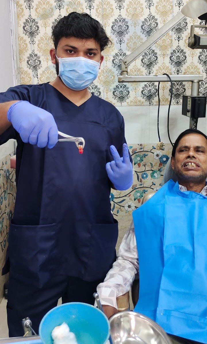 A person in a blue shirt with the logo 'Angels for Humanity' provides dental care to a young patient lying on a dental chair. The setting appears to be a makeshift dental clinic with blue medical equipment and supplies on tables. Other team members in similar attire are visible in the background, some attending to other patients.